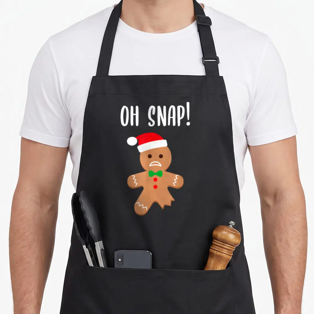 Outfit gingerbread apron - Cartoon gingerbread man in Santa hat and green bow tie below white OH SNAP text — shown on a black cotton canvas apron against a white background