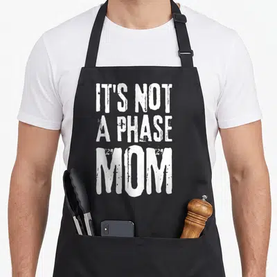 Phase lifestyle apron - White distressed stacked text reading IT'S NOT A PHASE MOM — shown on a black cotton canvas apron against a white background