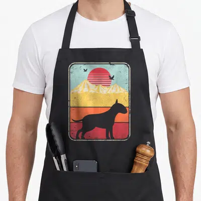 Terrier bull apron - Grey dog silhouette standing before yellow mountains and a red striped sun against a light blue sky — shown on a black cotton canvas apron against a white background