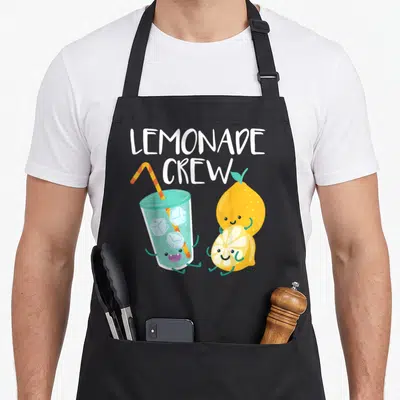 Hustle lemonade apron - Cartoon lemonade glass and smiling lemons with LEMONADE CREW text — shown on a black cotton canvas apron against a white background