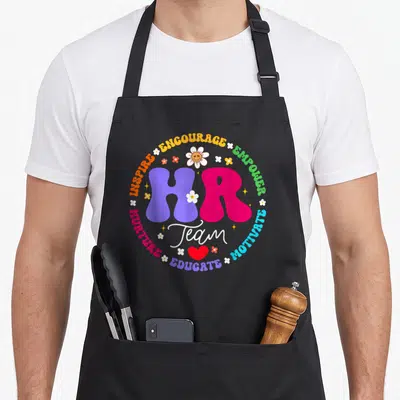 Human employee aprons - Colorful HR Team typography surrounded by encouraging verbs, flowers, stars, and a red heart — shown on a black cotton canvas apron against a white background
