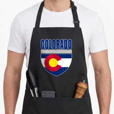 Colorado patriotic apron - Shield design with COLORADO text above blue white red and yellow flag elements — shown on a black cotton canvas apron against a white background