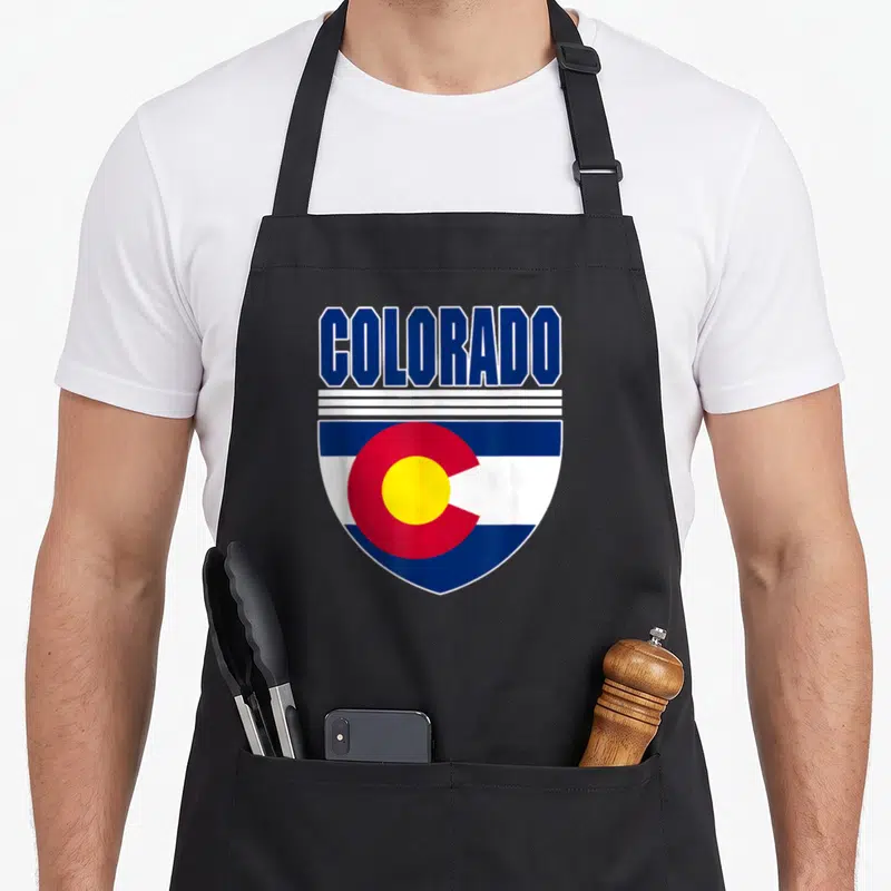 Colorado patriotic apron - Shield design with COLORADO text above blue white red and yellow flag elements — shown on a black cotton canvas apron against a white background