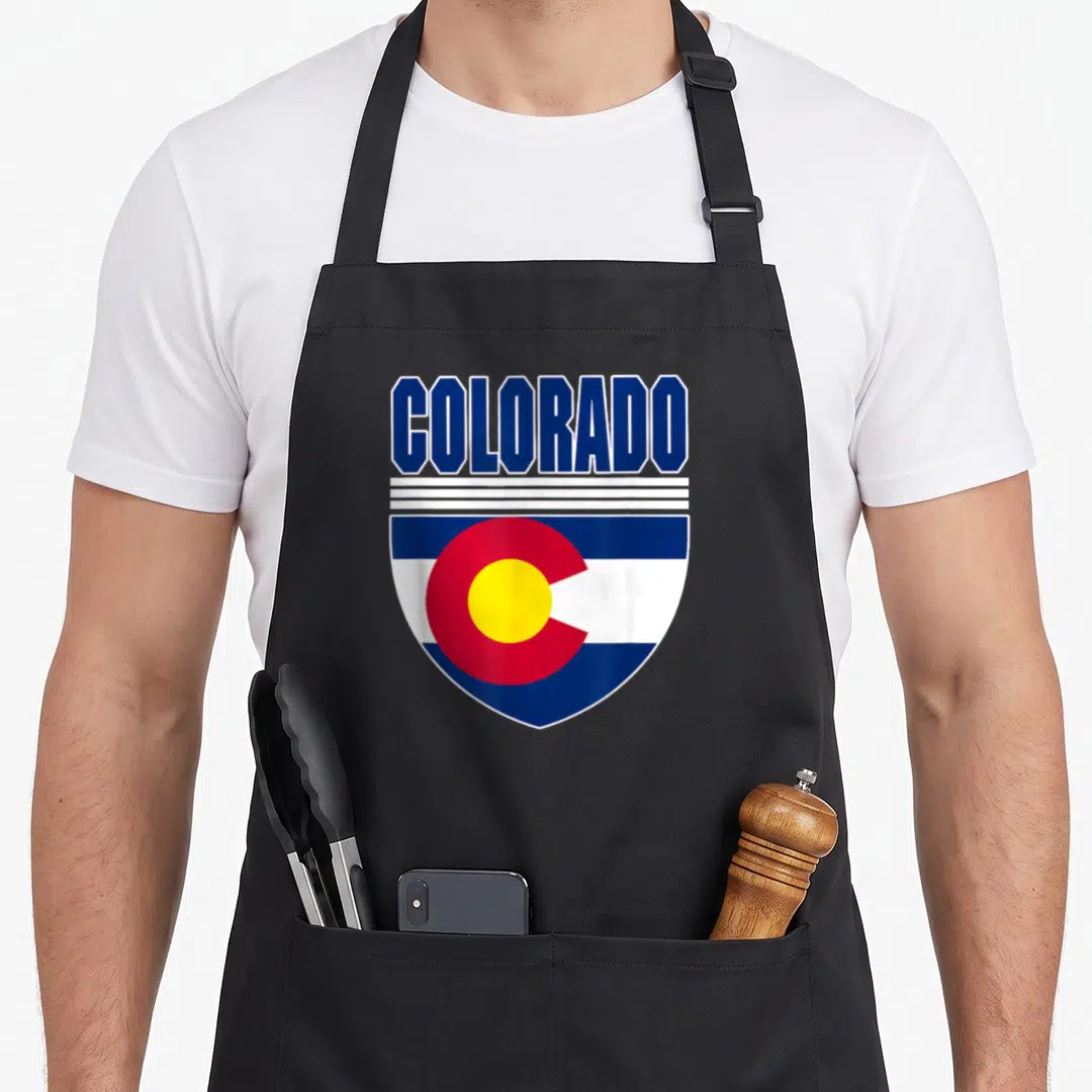 Colorado patriotic apron - Shield design with COLORADO text above blue white red and yellow flag elements — shown on a black cotton canvas apron against a white background