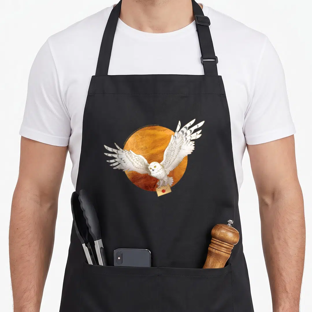 Hedwig potter apron - Realistic white owl flying before orange circle carrying tan envelope in talons — shown on a black cotton canvas apron against a white background