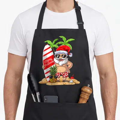 Surfing christmas santa apron - Cartoon Santa holding Merry Christmas surfboard on sand with sunglass-wearing pineapple — shown on a black cotton canvas apron against a white background
