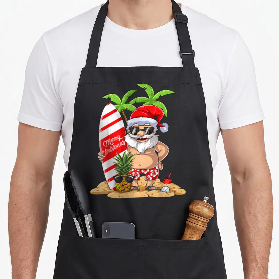 Surfing christmas santa apron - Cartoon Santa holding Merry Christmas surfboard on sand with sunglass-wearing pineapple — shown on a black cotton canvas apron against a white background