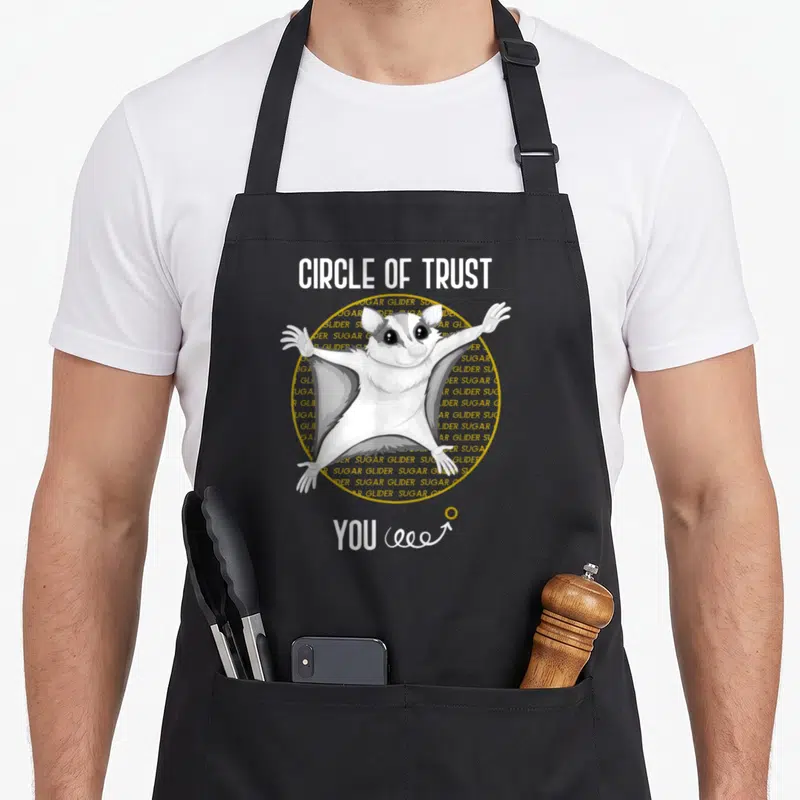 Glider pet sugar apron - Cartoon sugar glider in yellow circle with text CIRCLE OF TRUST YOU and spiral arrow — shown on a black cotton canvas apron against a white background