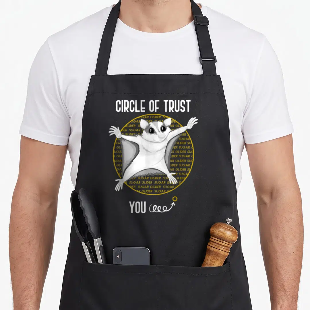 Glider pet sugar apron - Cartoon sugar glider in yellow circle with text CIRCLE OF TRUST YOU and spiral arrow — shown on a black cotton canvas apron against a white background