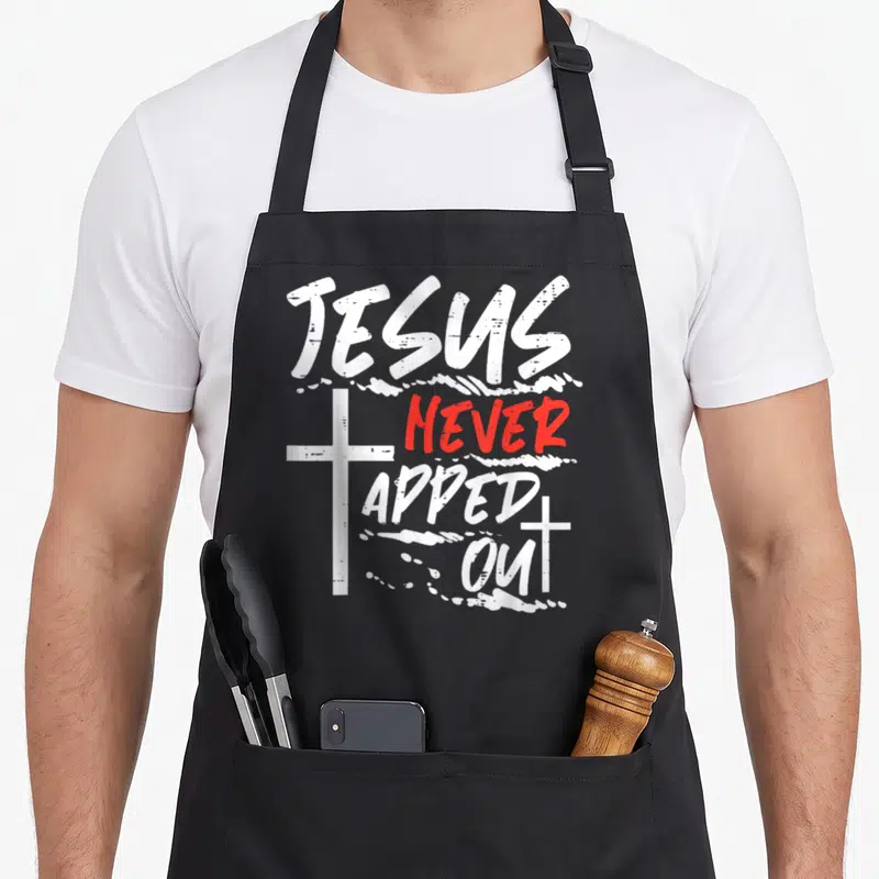Jesus wrestling apron - White and red text reading Jesus Never Tapped Out flanked by two distressed crosses — shown on a black cotton canvas apron against a white background