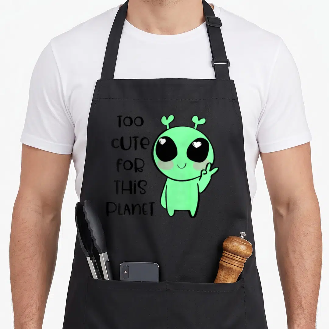 Alien kawaii apron - Light green alien character with heart eyes and stacked TOO CUTE FOR THIS PLANET text — shown on a black cotton canvas apron against a white background