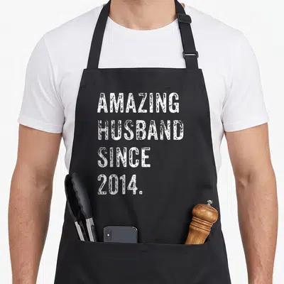 Amazing husband wedding aprons - White distressed text AMAZING HUSBAND SINCE 2014 stacked on black background — shown on a black cotton canvas apron against a white background