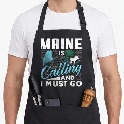 Calling maine girl apron - Distressed text reading MAINE IS Calling AND I MUST GO with moose and state outline — shown on a black cotton canvas apron against a white background