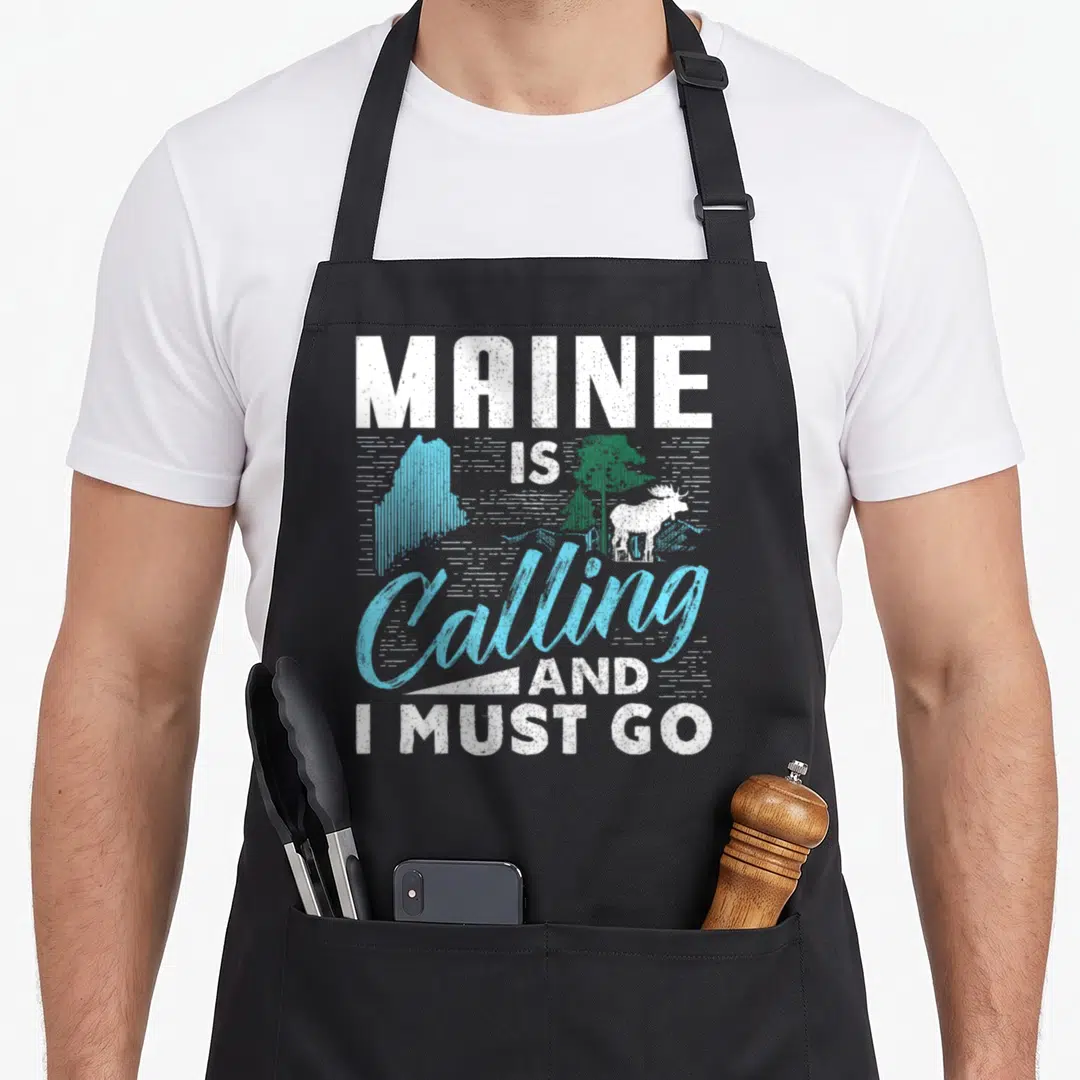 Calling maine girl apron - Distressed text reading MAINE IS Calling AND I MUST GO with moose and state outline — shown on a black cotton canvas apron against a white background