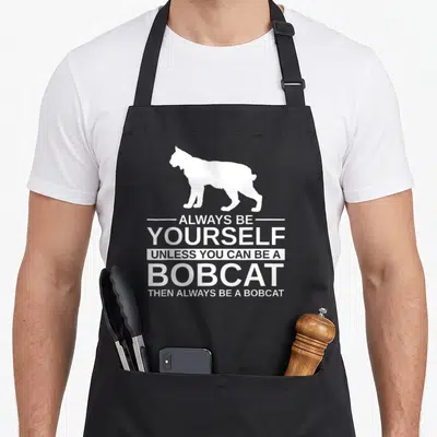 Alway kitty aprons - White bobcat silhouette above five lines of stacked white text reading Always Be Yourself Unless — shown on a black cotton canvas apron against a white background