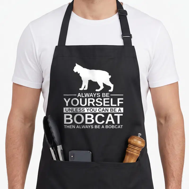Alway kitty aprons - White bobcat silhouette above five lines of stacked white text reading Always Be Yourself Unless — shown on a black cotton canvas apron against a white background