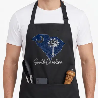 Palmetto pride apron - Navy South Carolina map with white moon palm tree and script text in distressed style — shown on a black cotton canvas apron against a white background