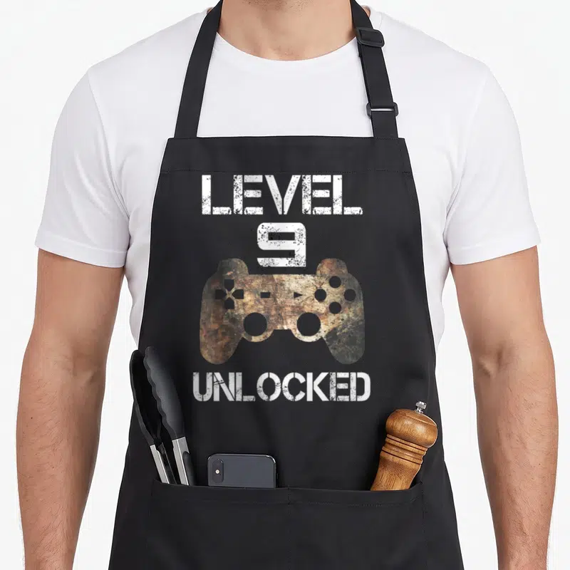 Unlocked 9th gamer birthday apron - Distressed white text level 9 unlocked surrounding a brown game controller — shown on a black cotton canvas apron against a white background