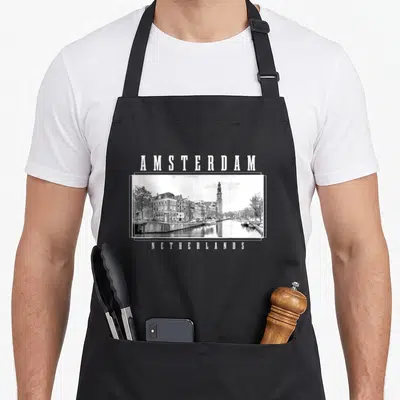 Amsterdam netherlands apron - White AMSTERDAM text above grey black framed canal photo, NETHERLANDS text below — shown on a black cotton canvas apron against a white background