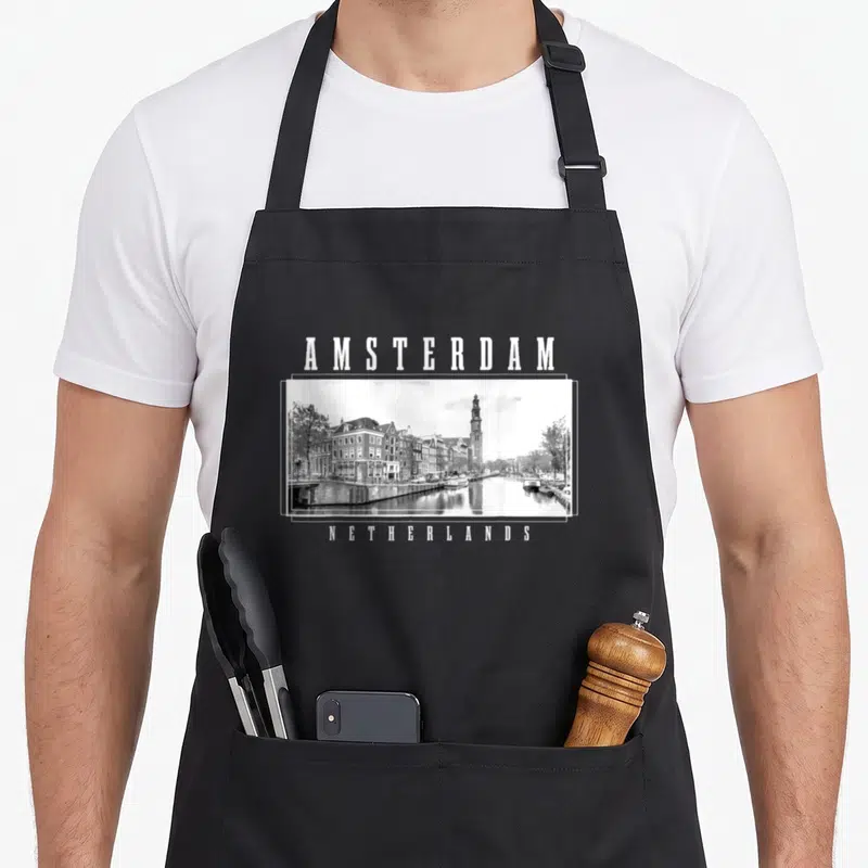Amsterdam netherlands apron - White AMSTERDAM text above grey black framed canal photo, NETHERLANDS text below — shown on a black cotton canvas apron against a white background