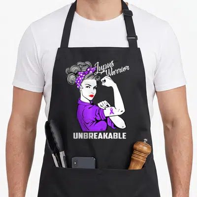 Warrior lupus awareness apron - Cartoon woman flexing arm with purple ribbon and Lupus Warrior UNBREAKABLE text — shown on a black cotton canvas apron against a white background