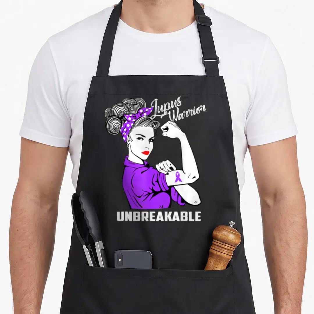 Warrior lupus awareness apron - Cartoon woman flexing arm with purple ribbon and Lupus Warrior UNBREAKABLE text — shown on a black cotton canvas apron against a white background