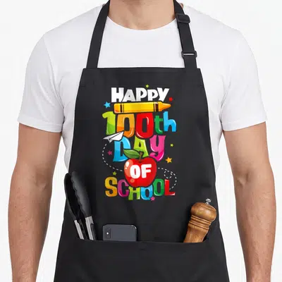 100th teacher apron - Stacked HAPPY 100th Day OF SCHOOL text with crayon, plane, apple, and stars in red yellow blue — shown on a black cotton canvas apron against a white background