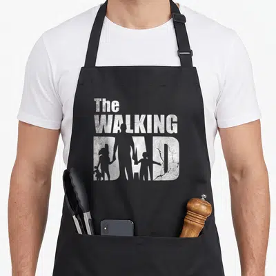 Walking father day apron - Distressed white text reading The Walking Dad with father and two child silhouettes cut — shown on a black cotton canvas apron against a white background