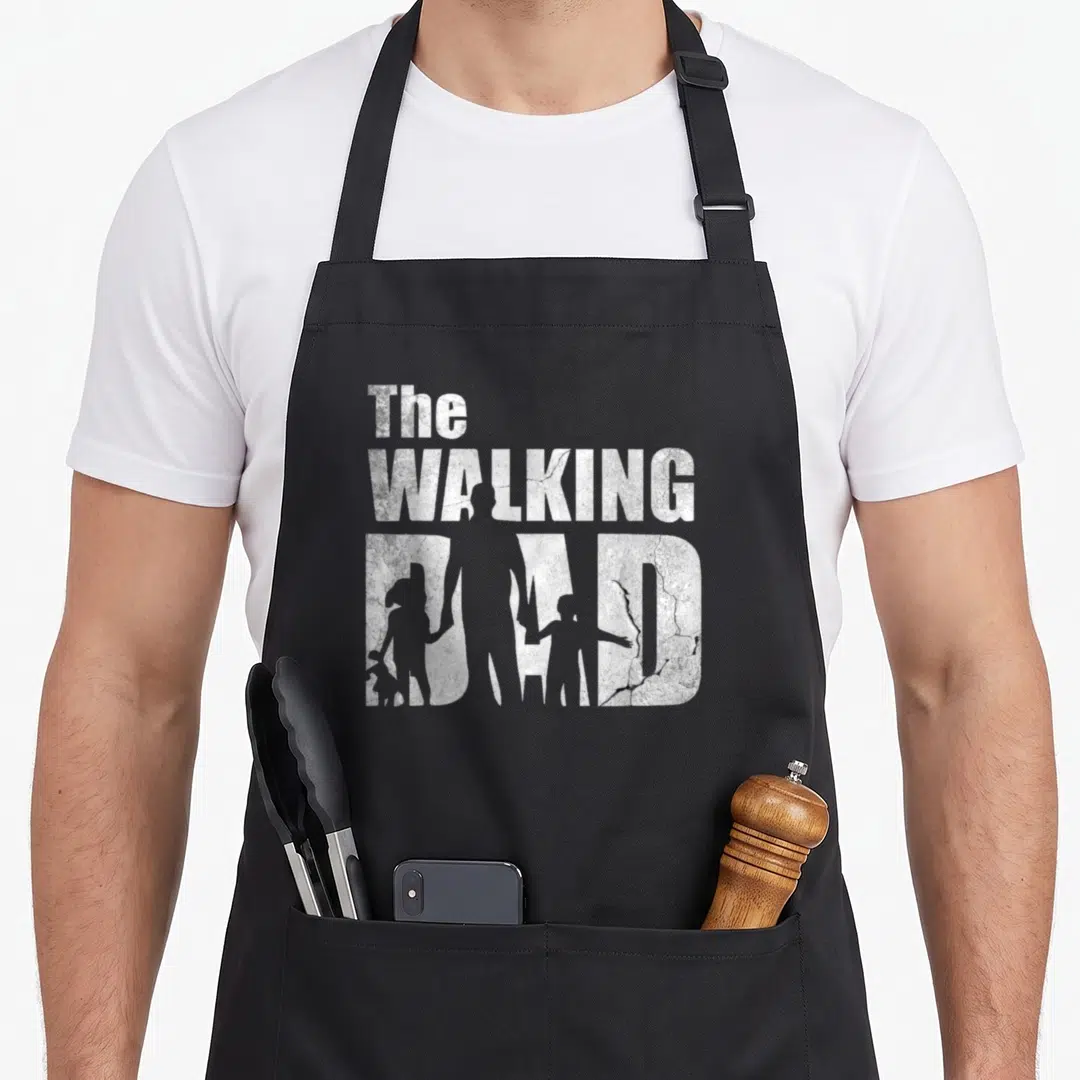 Walking father day apron - Distressed white text reading The Walking Dad with father and two child silhouettes cut — shown on a black cotton canvas apron against a white background