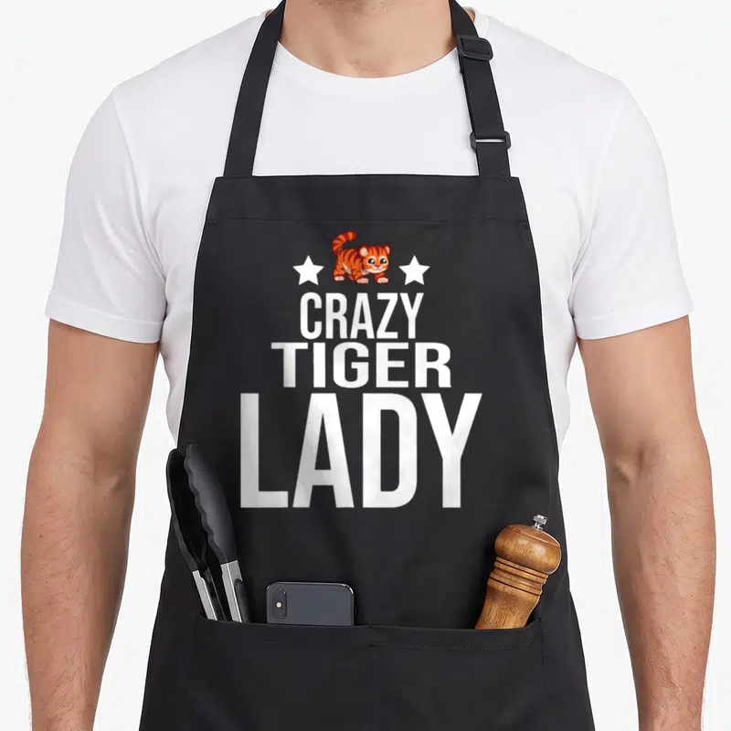Cute tiger animal apron - Cartoon tiger between two stars above stacked white text reading crazy tiger lady — shown on a black cotton canvas apron against a white background