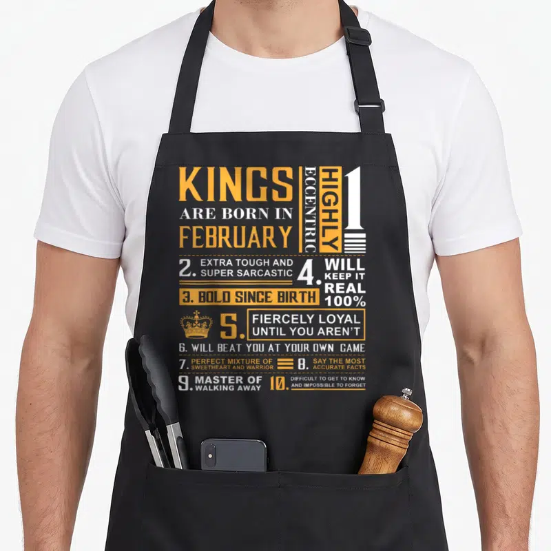 February king apron - Yellow and white typography grid listing ten eccentric traits of kings born in February including — shown on a black cotton canvas apron against a white background