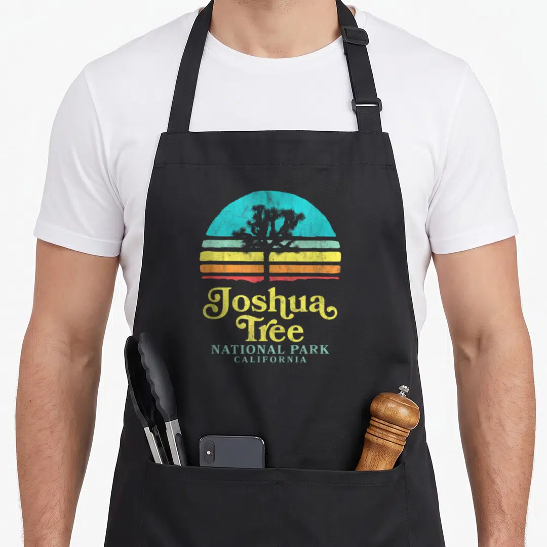 National park tree apron - Retro Joshua Tree silhouette over striped sun with stacked text Joshua Tree National Park — shown on a black cotton canvas apron against a white background