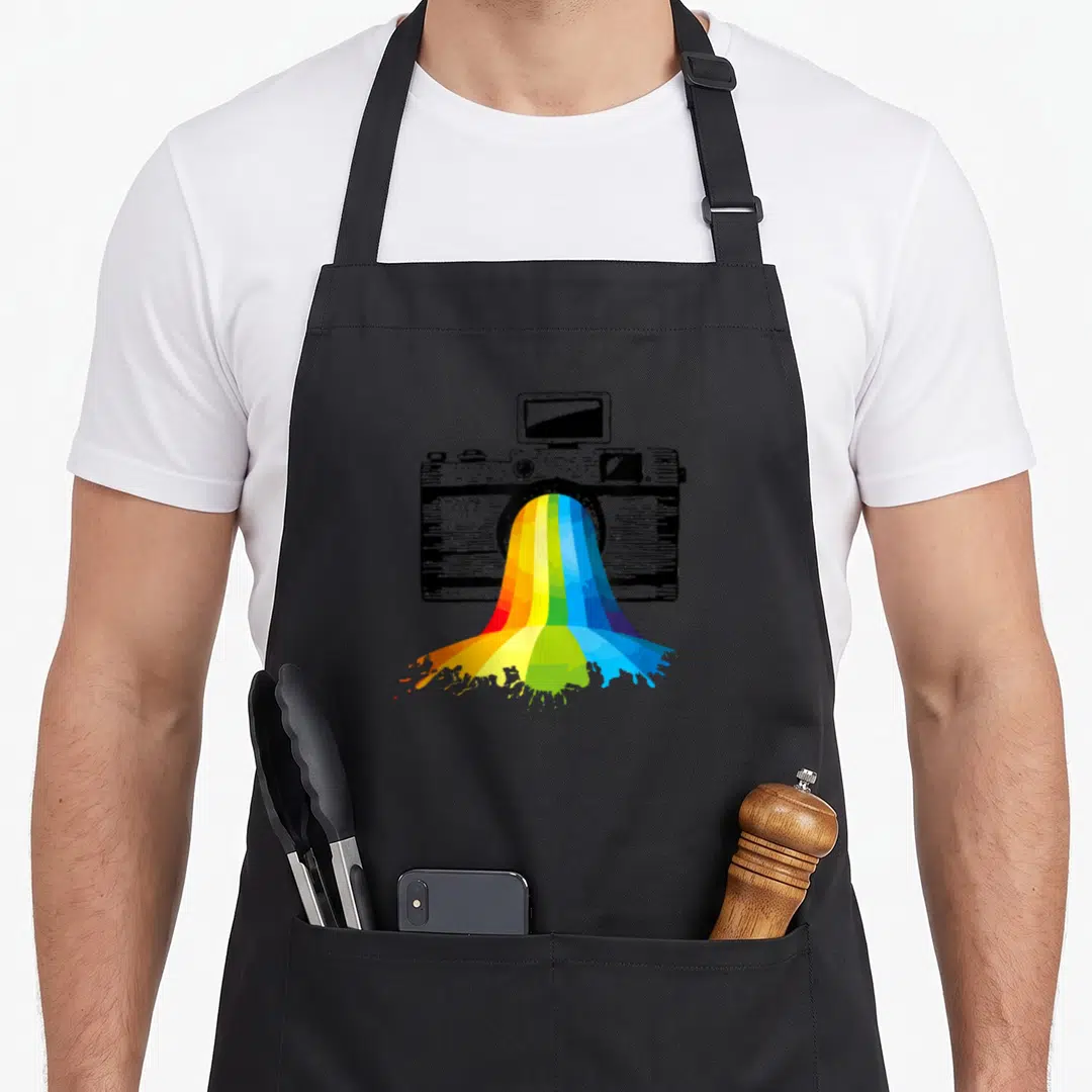 Photography rainbow apron - Black and white camera sketch with rainbow paint splashing from lens in red orange yellow — shown on a black cotton canvas apron against a white background