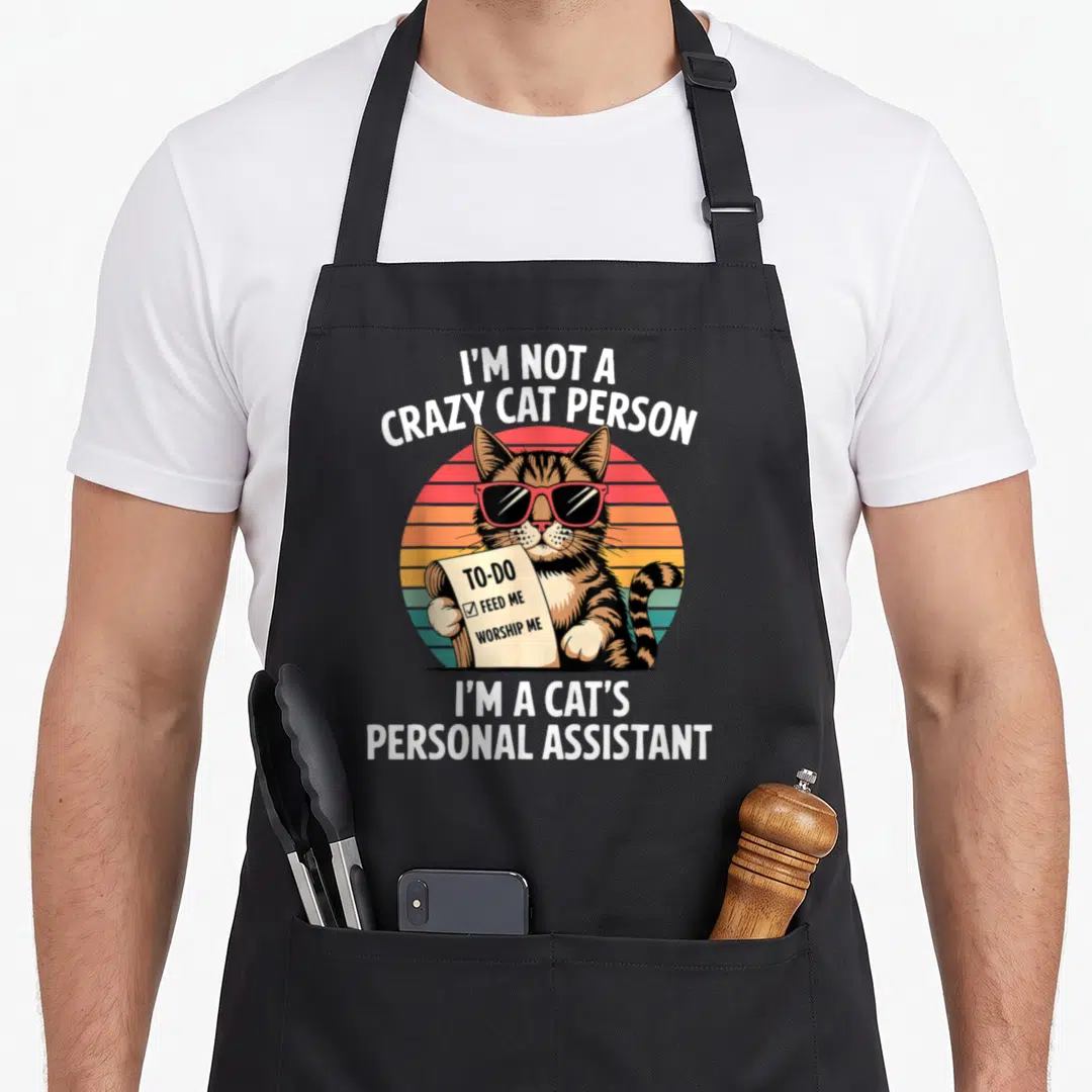 Cat apron for hosting - Retro cartoon cat in sunglasses holding a to-do list with text about being a personal assistant — shown on a black cotton canvas apron against a white background