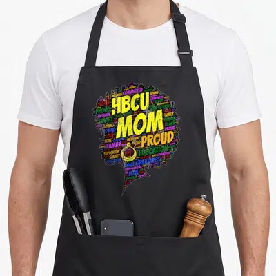 Hbcu graduation apron - HBCU MOM PROUD yellow text over colorful word cloud woman silhouette — shown on a black cotton canvas apron against a white background