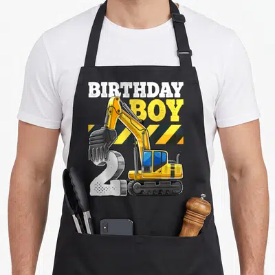 Excavator 2nd apron - Yellow excavator digging white number 2 with BIRTHDAY BOY text and hazard stripes — shown on a black cotton canvas apron against a white background