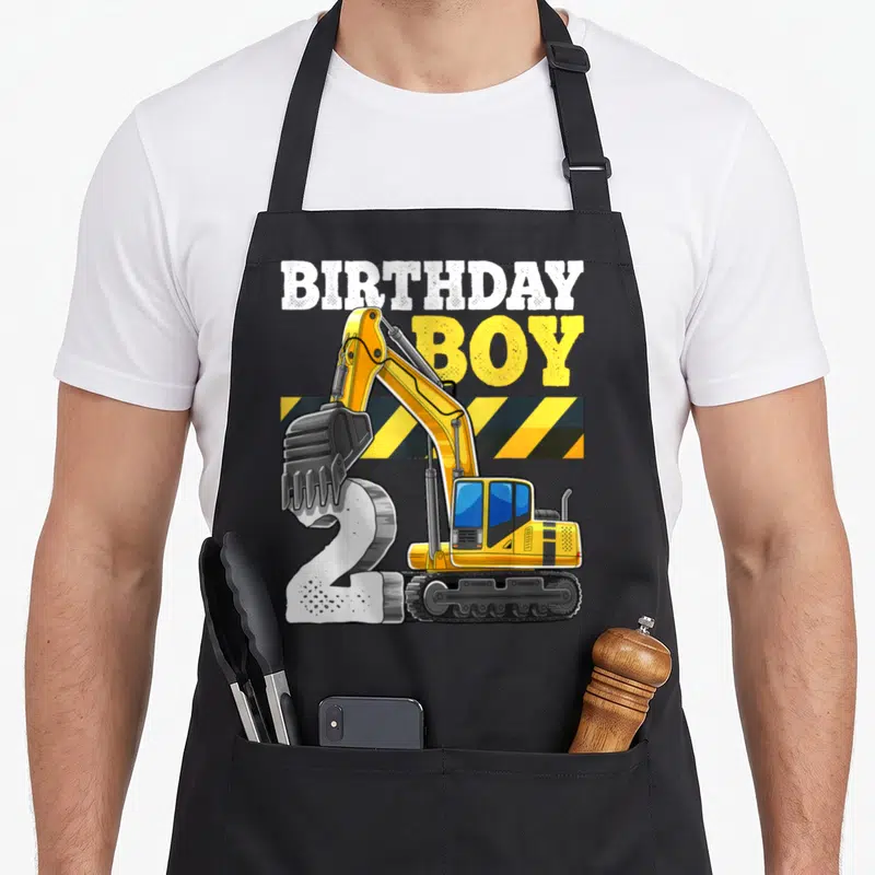 Excavator 2nd apron - Yellow excavator digging white number 2 with BIRTHDAY BOY text and hazard stripes — shown on a black cotton canvas apron against a white background