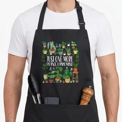 Promise gardener apron - Just One More Plant I Promise text surrounded by cartoon houseplants in pots — shown on a black cotton canvas apron against a white background