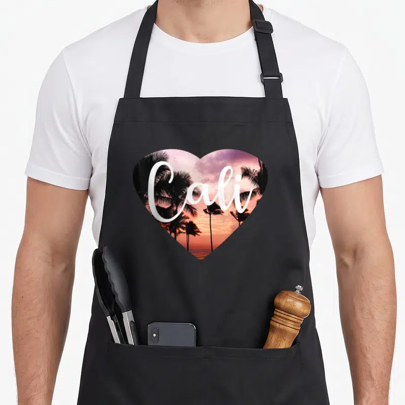 Funny cali california apron - Heart shaped beach sunset photo with palm trees and white Cali text in orange pink black — shown on a black cotton canvas apron against a white background