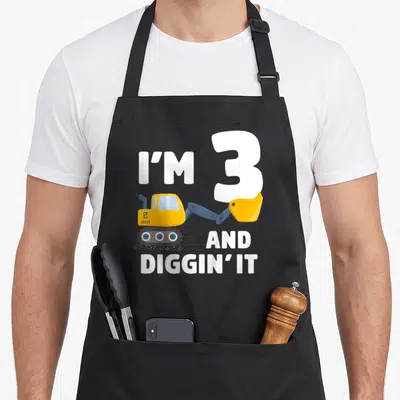 Birthday truck apron - I'M 3 AND DIGGIN' IT text with yellow excavator illustration — shown on a black cotton canvas apron against a white background