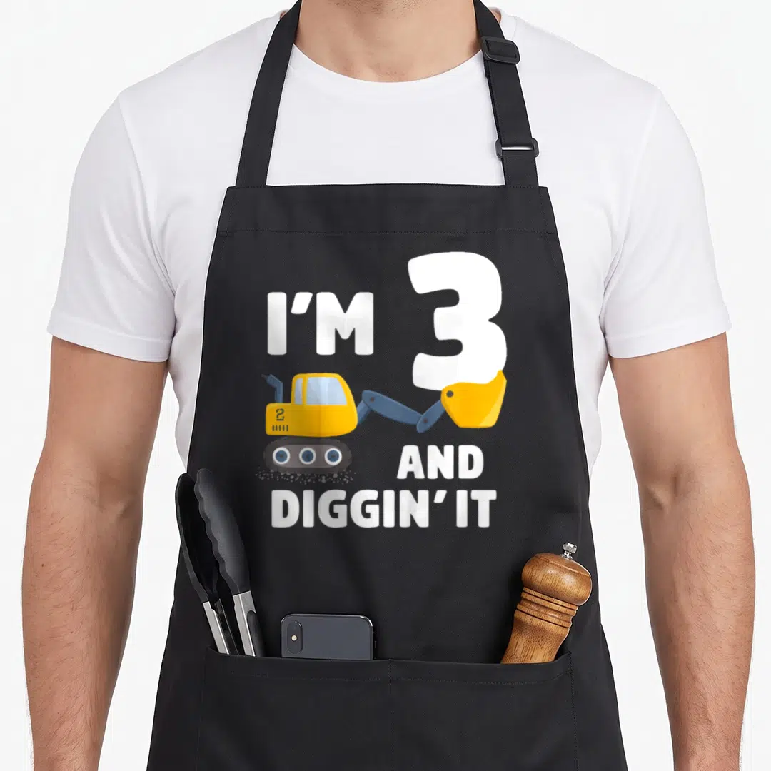 Birthday truck apron - I'M 3 AND DIGGIN' IT text with yellow excavator illustration — shown on a black cotton canvas apron against a white background