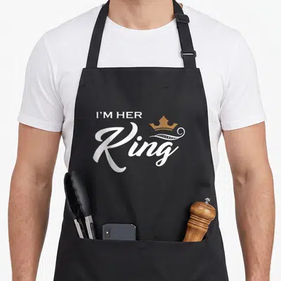 King wife apron - White text I'M HER Ring with gold crown and decorative swirl above the letter R — shown on a black cotton canvas apron against a white background
