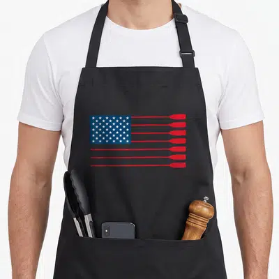 Rowing american flag apron - Stylized US flag with red oars replacing stripes and white stars on blue field — shown on a black cotton canvas apron against a white background
