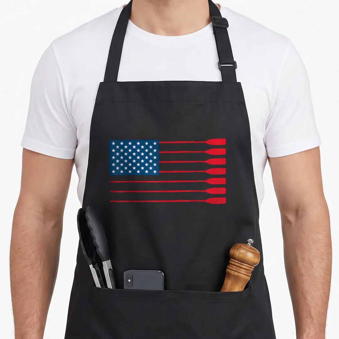 Rowing american flag apron - Stylized US flag with red oars replacing stripes and white stars on blue field — shown on a black cotton canvas apron against a white background