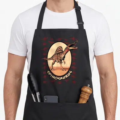 Cute spinosaurus dinosaur apron - Realistic spinosaurus in oval with red silhouettes and arched SPINOSAURUS text — shown on a black cotton canvas apron against a white background