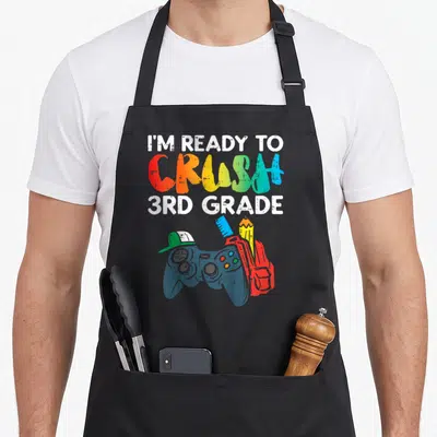 Crush day grade apron - Rainbow text I'm Ready To Crush 3rd Grade above game controller and backpack illustration — shown on a black cotton canvas apron against a white background