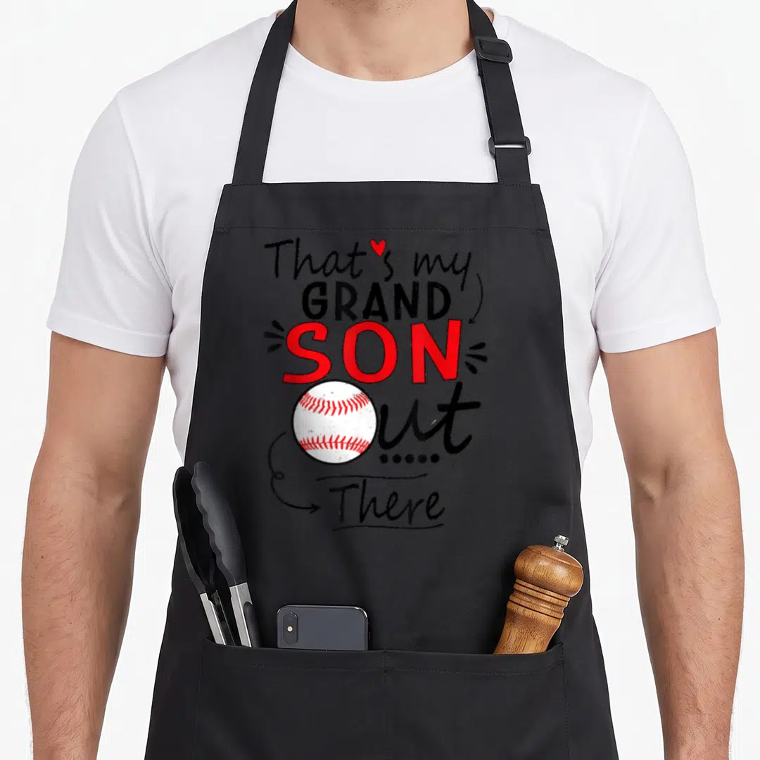 Cute grandson grandma apron - Stacked text That's my GRAND SON Out There with baseball and heart in black red white — shown on a black cotton canvas apron against a white background