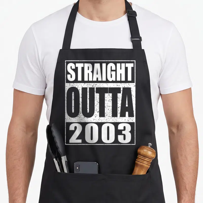 Outta 22nd birthday apron - Stacked white and grey distressed text reading STRAIGHT OUTTA 2003 inside a rectangle — shown on a black cotton canvas apron against a white background