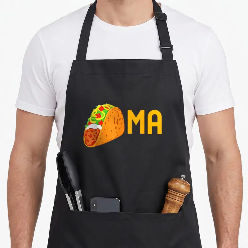 Tacoma taco apron - Cartoon taco next to yellow MA text with orange shell and colorful fillings — shown on a black cotton canvas apron against a white background