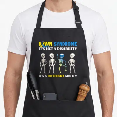 Month down syndrome awareness apron - Four cartoon skeletons, one rainbow-colored waving, with awareness text and ribbon — shown on a black cotton canvas apron against a white background